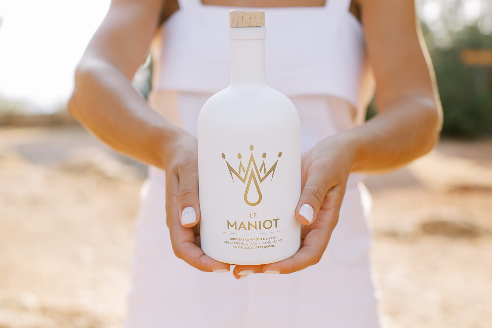 le-maniot-painted-glass-olive-oil-bottle-hands.jpg Hands present a matte white Le Maniot superior olive‑oil bottle with gold crown logo in sunlit Mani, Greece.